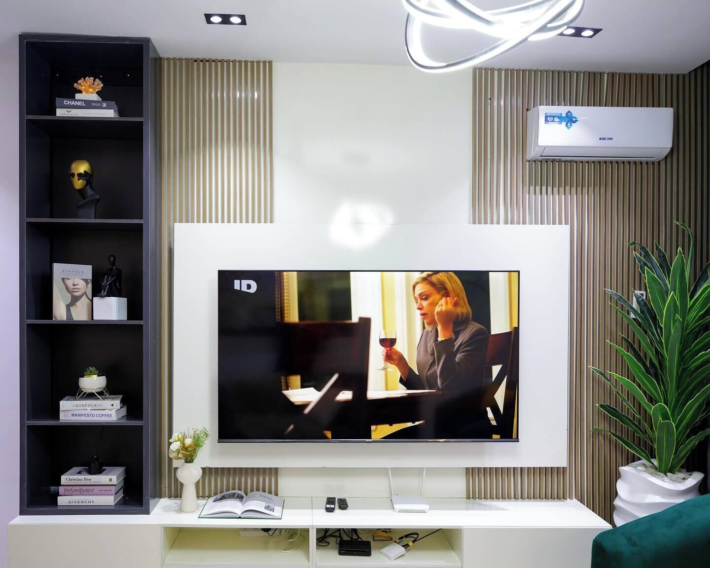 A modern living room with a flat-screen TV mounted on the wall, a bookshelf with various decorative items, a potted plant, and a wall-mounted air conditioning unit.
