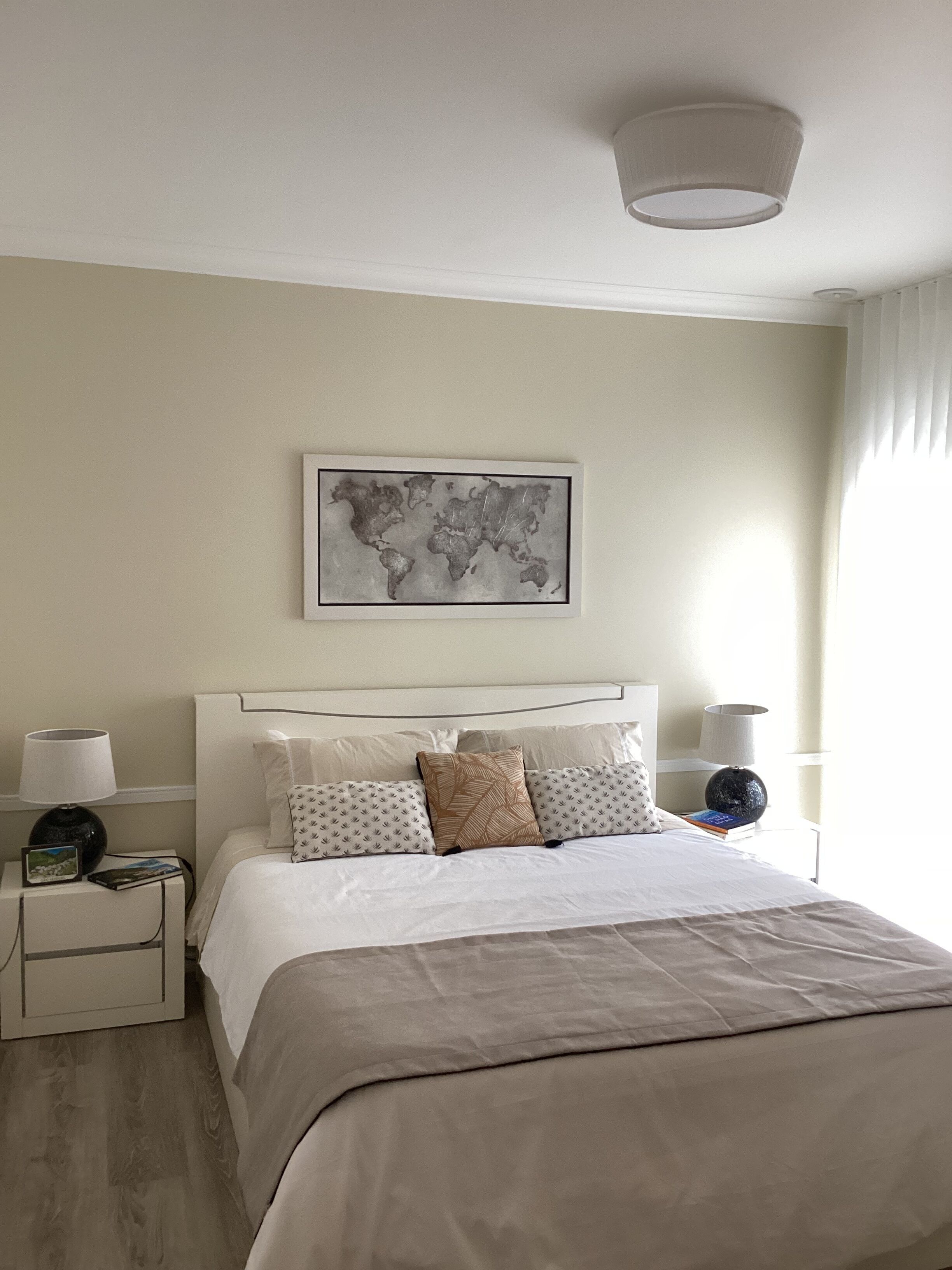 A neatly arranged bedroom with a large bed, two bedside tables, a world map artwork, and a ceiling light fixture.