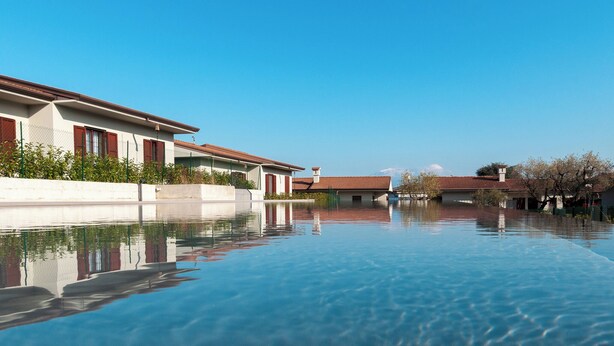 A clear swimming pool with a reflection of buildings, surrounded by a low wall and trees. - Aurora Lights Suite (Desenzano del Garda)