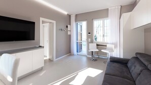 A modern living room with a grey sofa, a white chair, a flat-screen TV mounted on the wall, and a small table with a lamp. - Aurora Lights Suite (Desenzano del Garda)