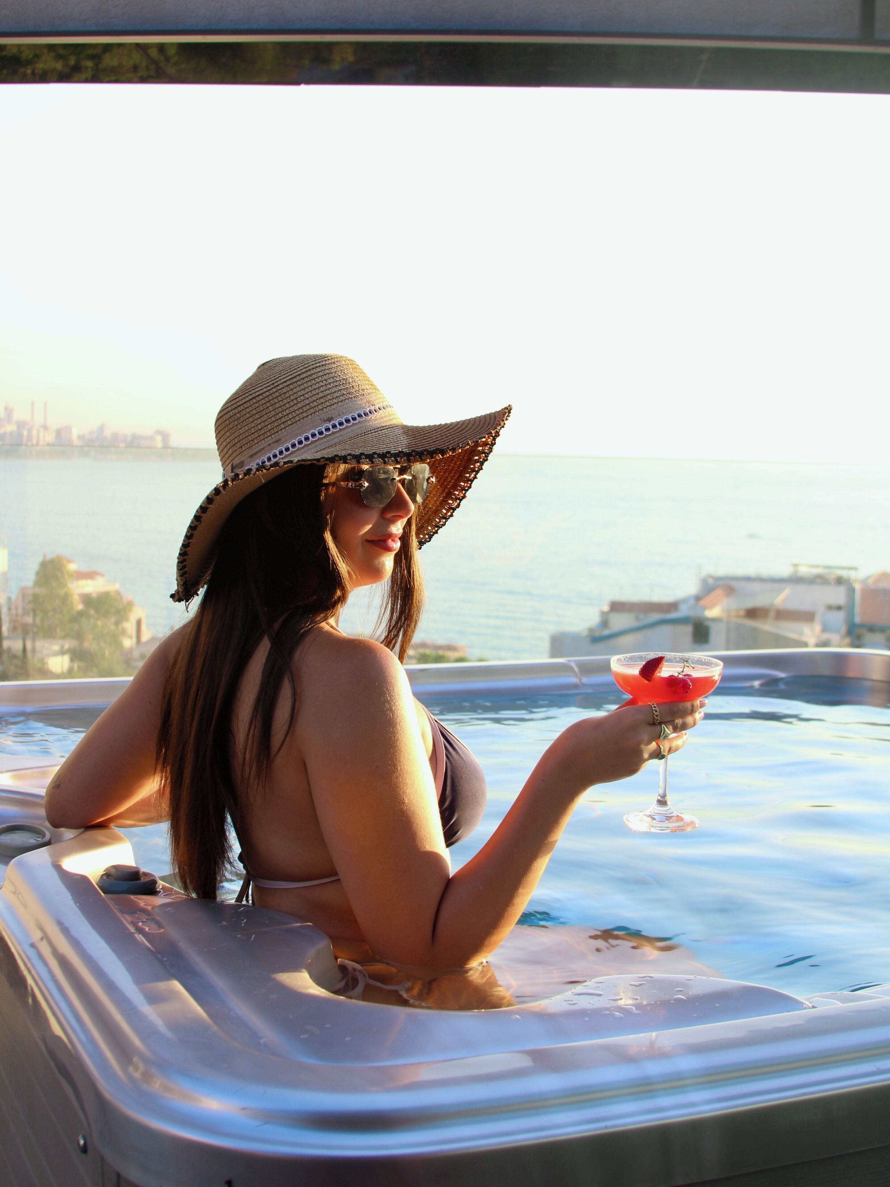 A person in a hot tub with a drink, overlooking a body of water and distant buildings.