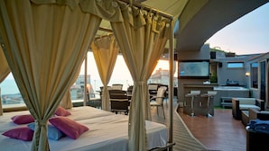 A balcony with a bed, chairs, a TV, and a grill. - Hotel Xperience (Ghazir)