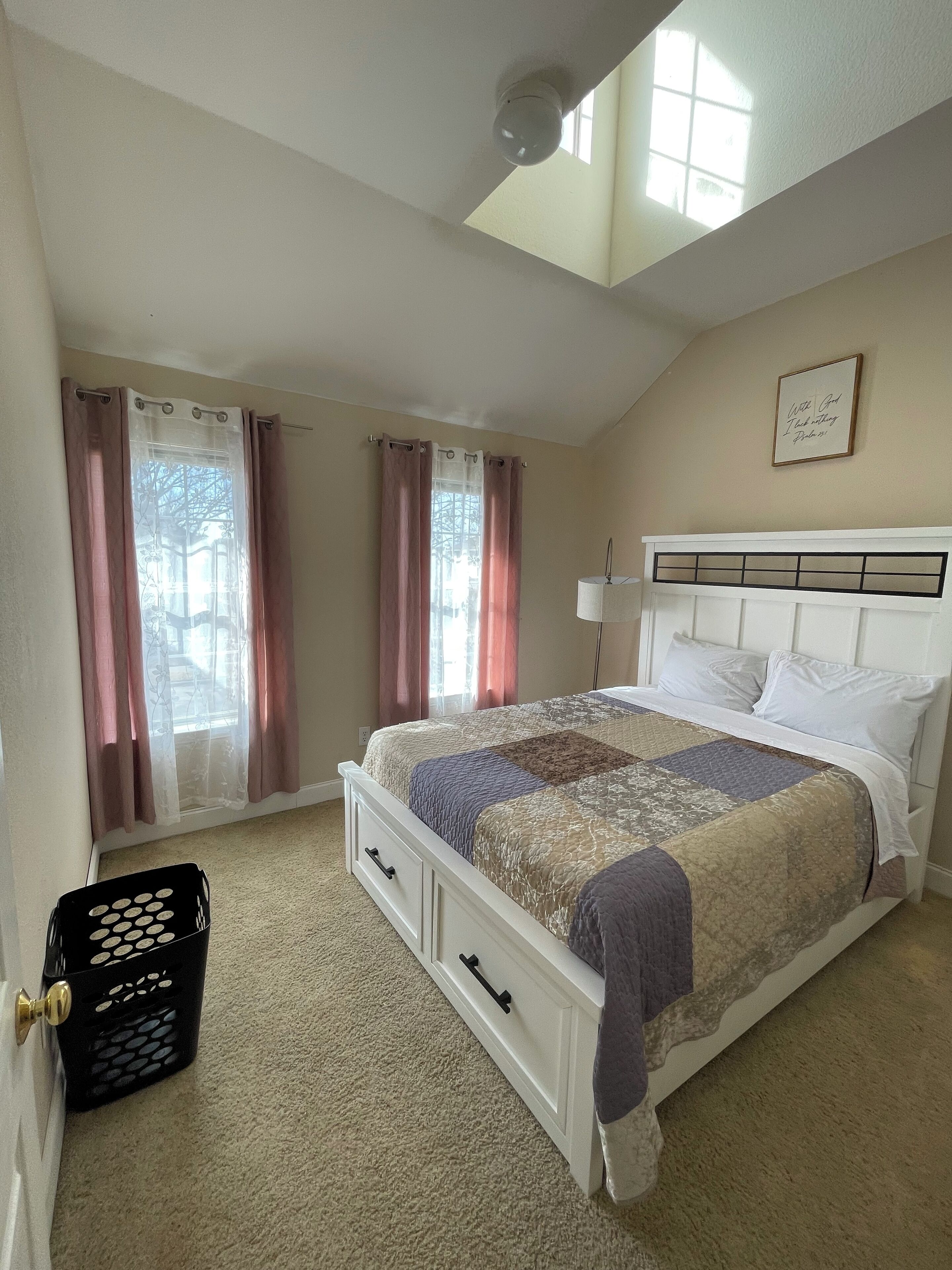 A bedroom with a bed, a ceiling fan, a window with curtains, a wall-mounted light, and a framed quote.