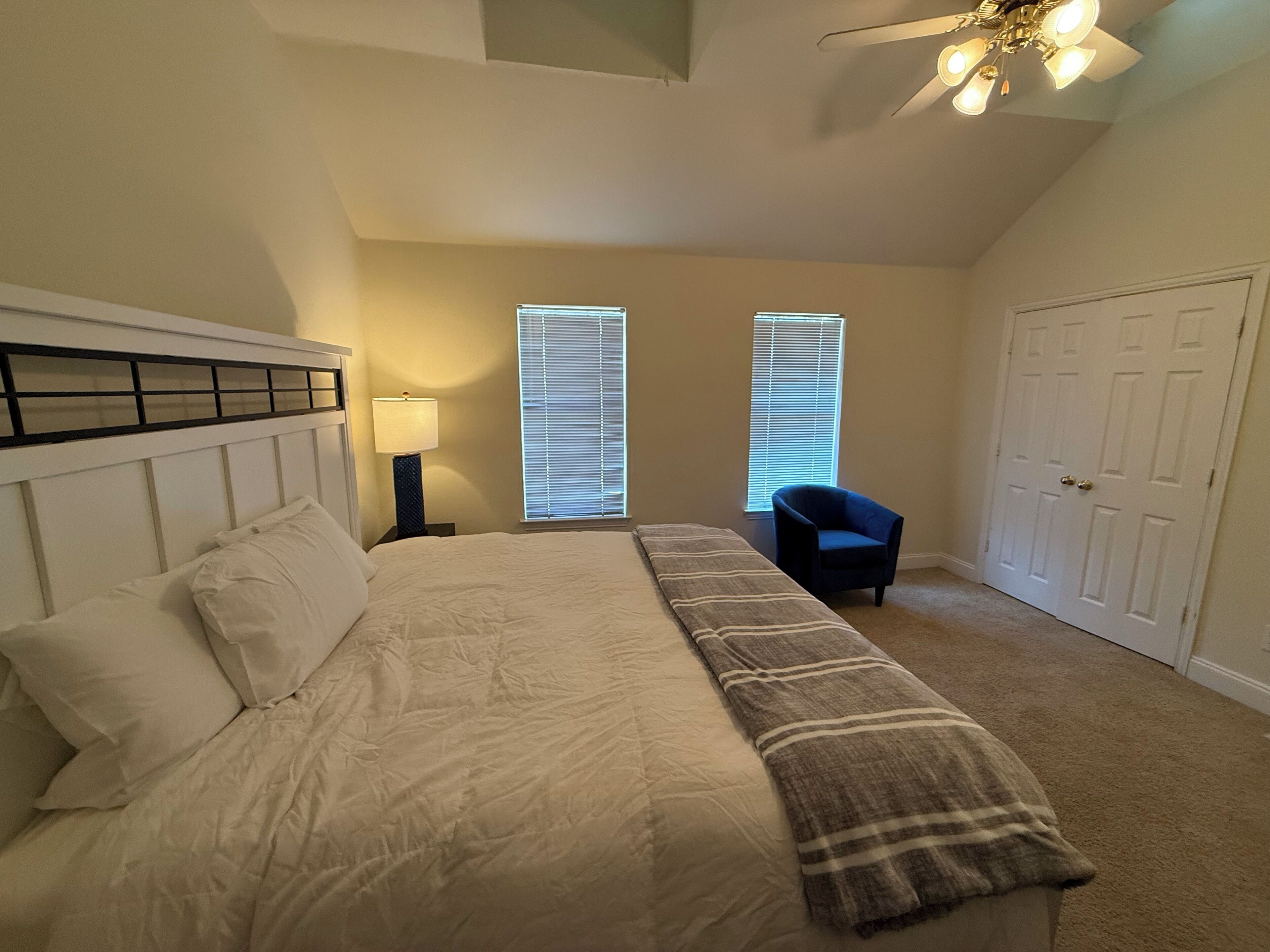 A bedroom with a bed, a nightstand, a chair, two windows with blinds, a ceiling fan, and a door.