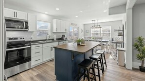 A modern kitchen with white cabinets, a central island, and a dining area with a table and chairs. - 4BR Channel House-Dock-Linda's Castaway Cottage (North Myrtle Beach)