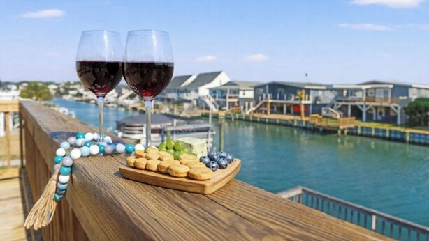 A wooden deck with two glasses of red wine, a beaded necklace, and a plate of snacks. - 4BR Channel House-Dock-Linda's Castaway Cottage (North Myrtle Beach)