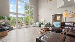 A spacious living room with a large window, a leather sofa, a television, and a dining area with outdoor views. - Mimosa Mornings Home for 12 (Eldon)