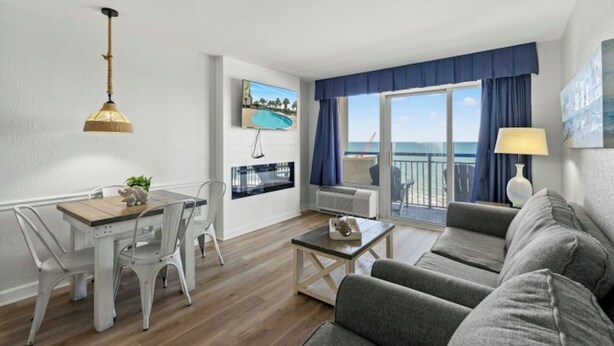 En moderne stue med sofa, spisebord med stole, fjernsyn ophængt på væggen og en balkon med havudsigt.