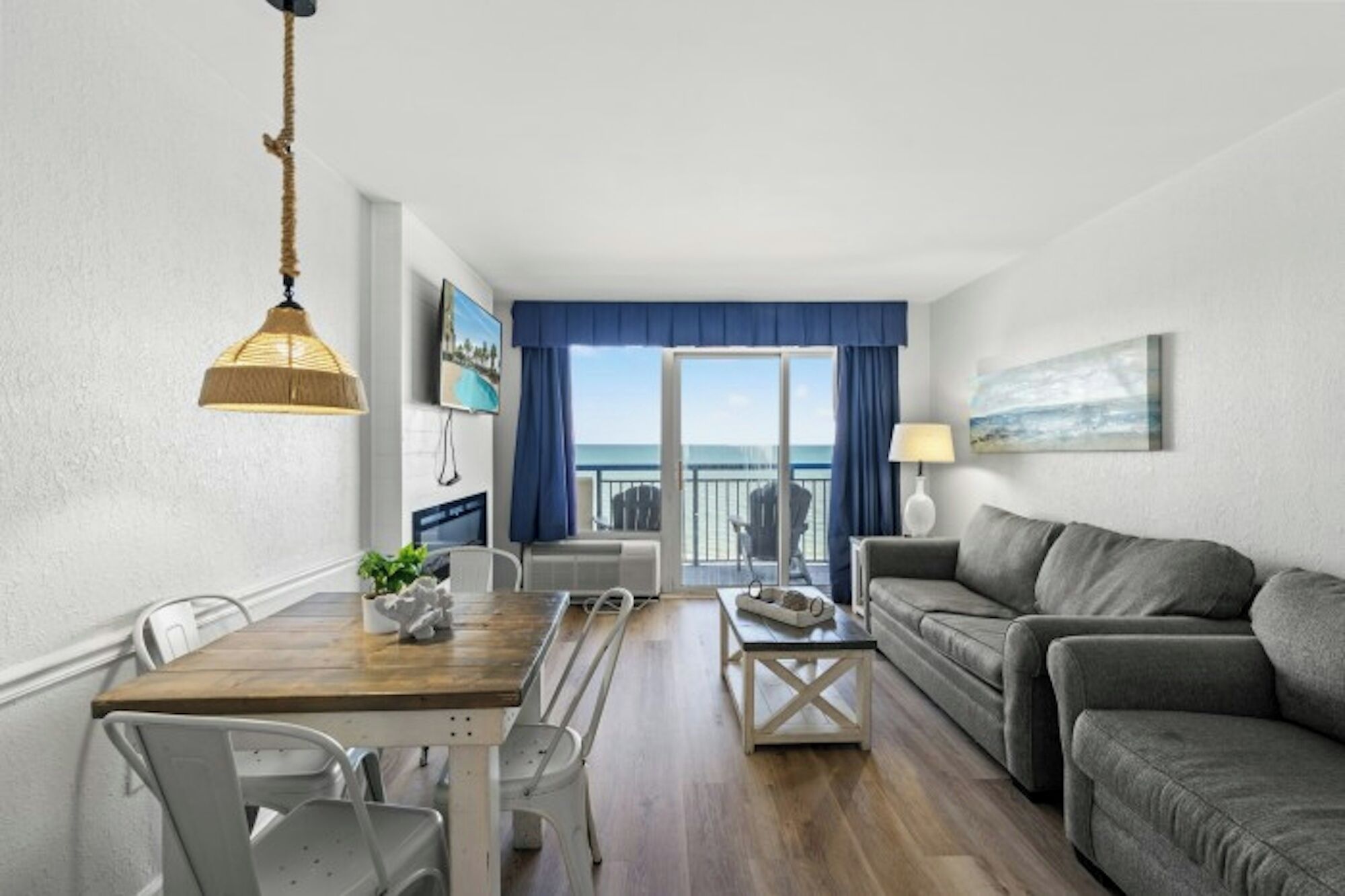A modern living room with a wooden dining table, grey sofa, and a balcony with a view of the sea.