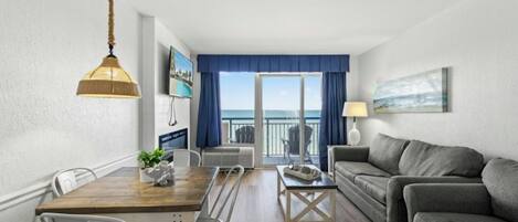 A modern living room with a wooden dining table, grey sofa, and a balcony with a view of the sea.