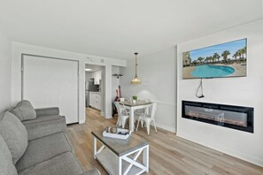 A modern living room with a grey sofa, a wooden coffee table, and a dining area with white chairs. A TV is mounted on the wall, and a fireplace is visible. - Boardwalk Beach Resort 939/940- Oceanfront 2BR (Myrtle Beach)