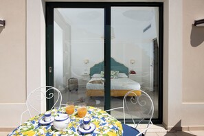 Una habitación con cama, una mesa con mantel floreado y un ventanal corredero que da a un balcón.