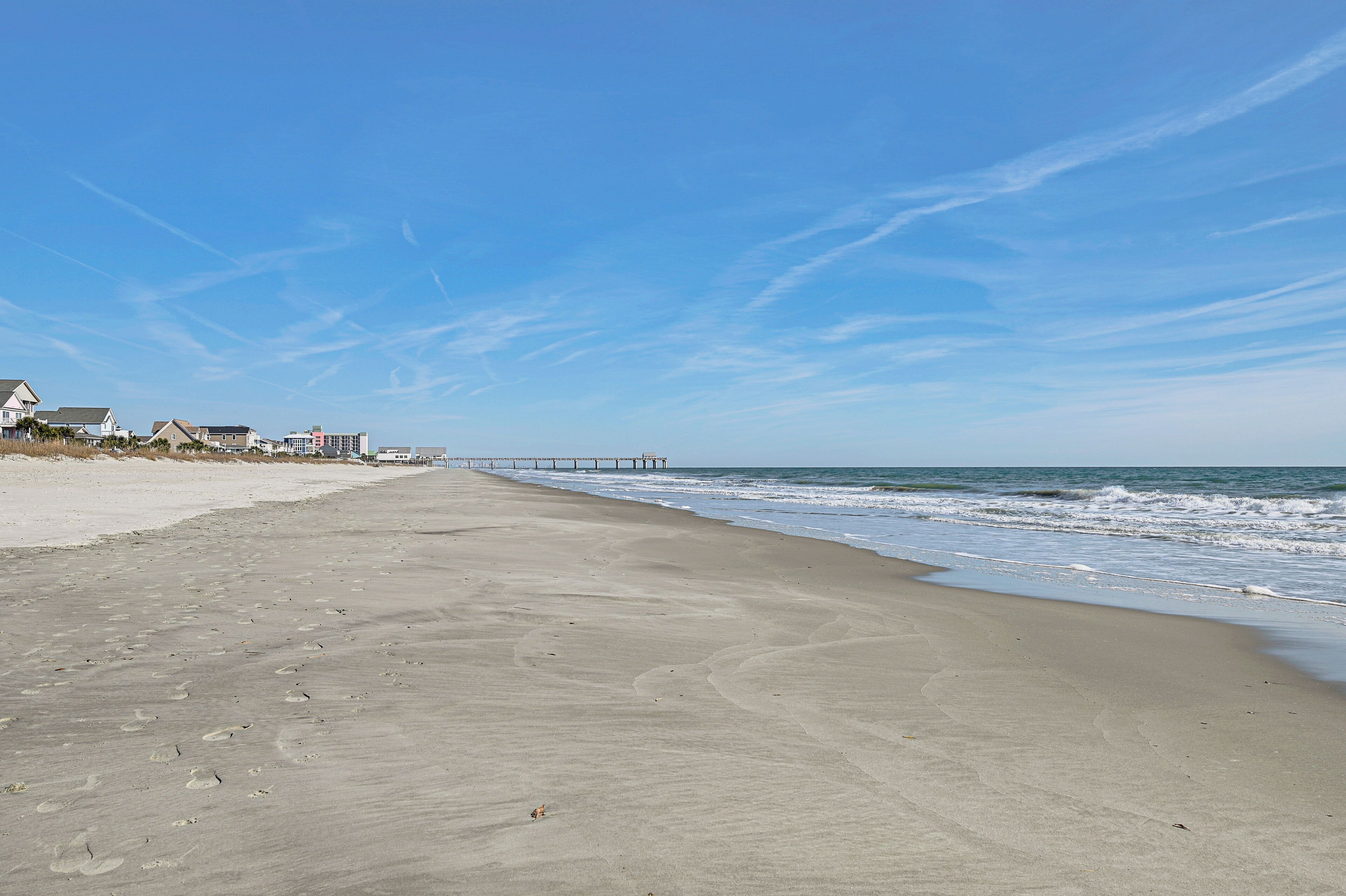 A beach with footprints, a pier, and houses in the background.