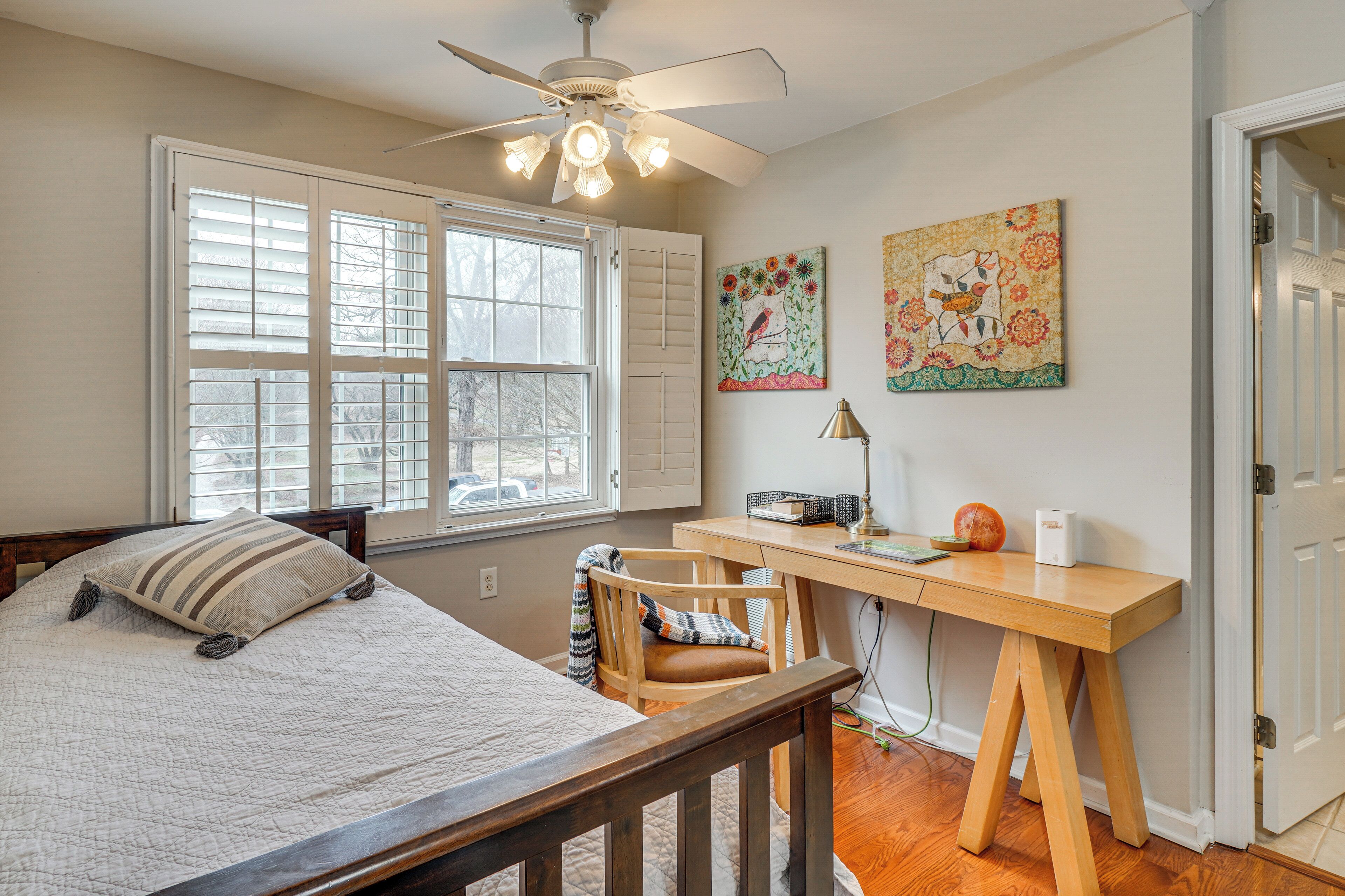A bedroom with a bed, a desk, and a chair. There is a window with blinds, a ceiling fan, and artwork on the wall.