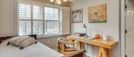 A bedroom with a bed, a desk, and a chair. There is a window with blinds, a ceiling fan, and artwork on the wall.