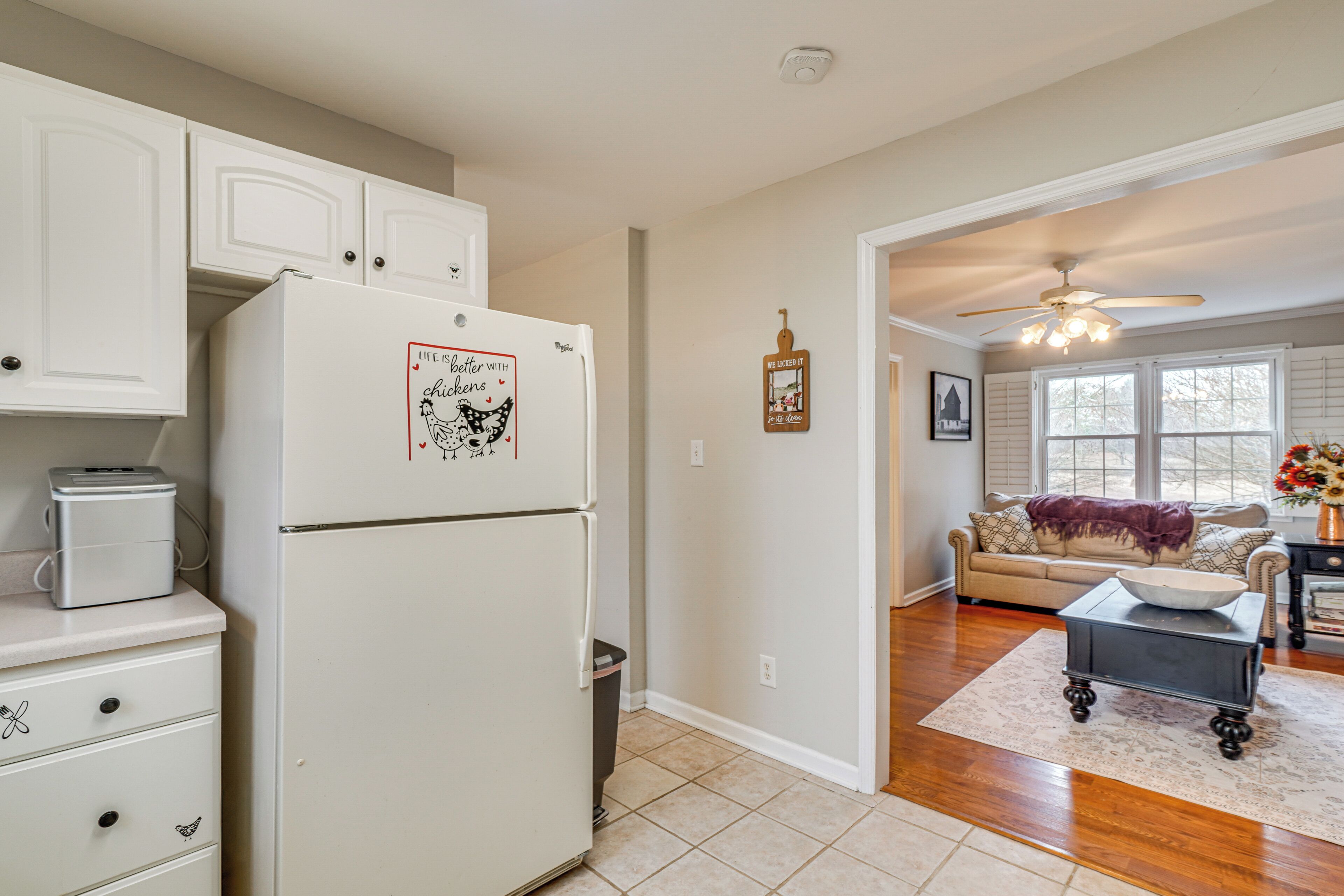 A kitchen with white cabinets, a refrigerator, a toaster, and a trash can. There is a dining area with a table, chairs, and a vase with flowers.