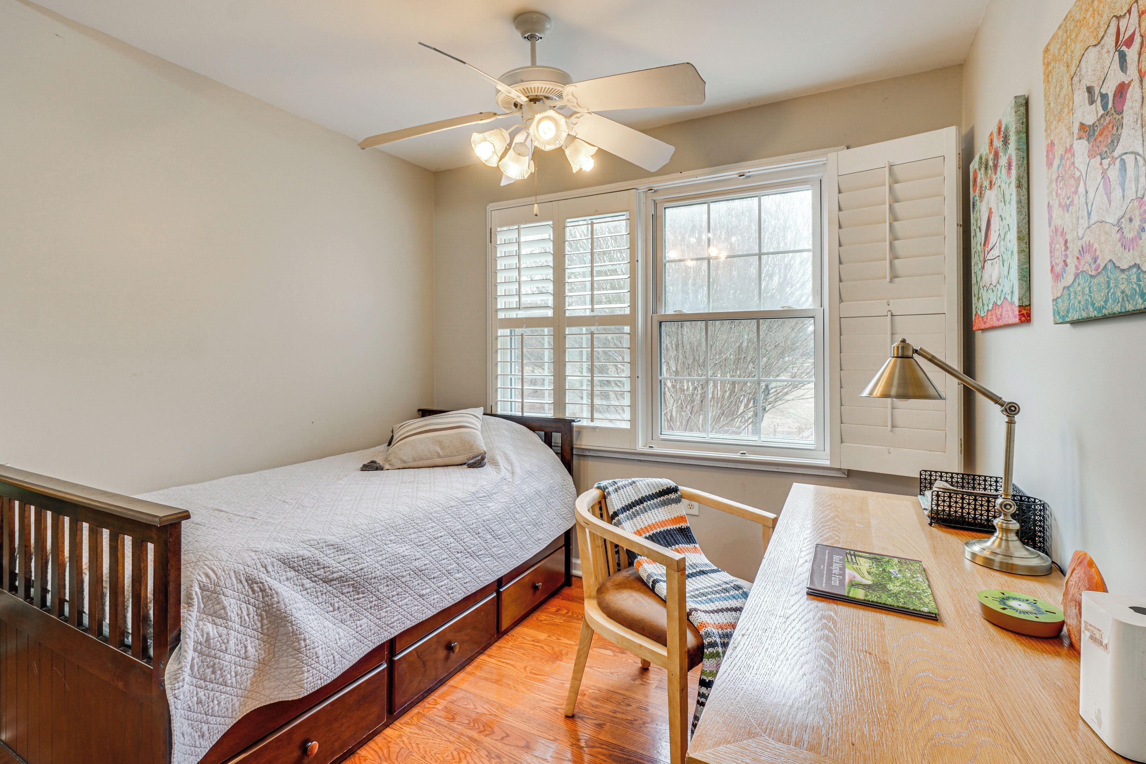 A bedroom with a bed, a desk, a chair, a ceiling fan, and a window with shutters.