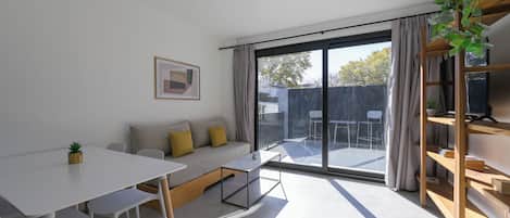 A modern living room with a sofa, a dining table, and a large sliding glass door leading to an outdoor area.
