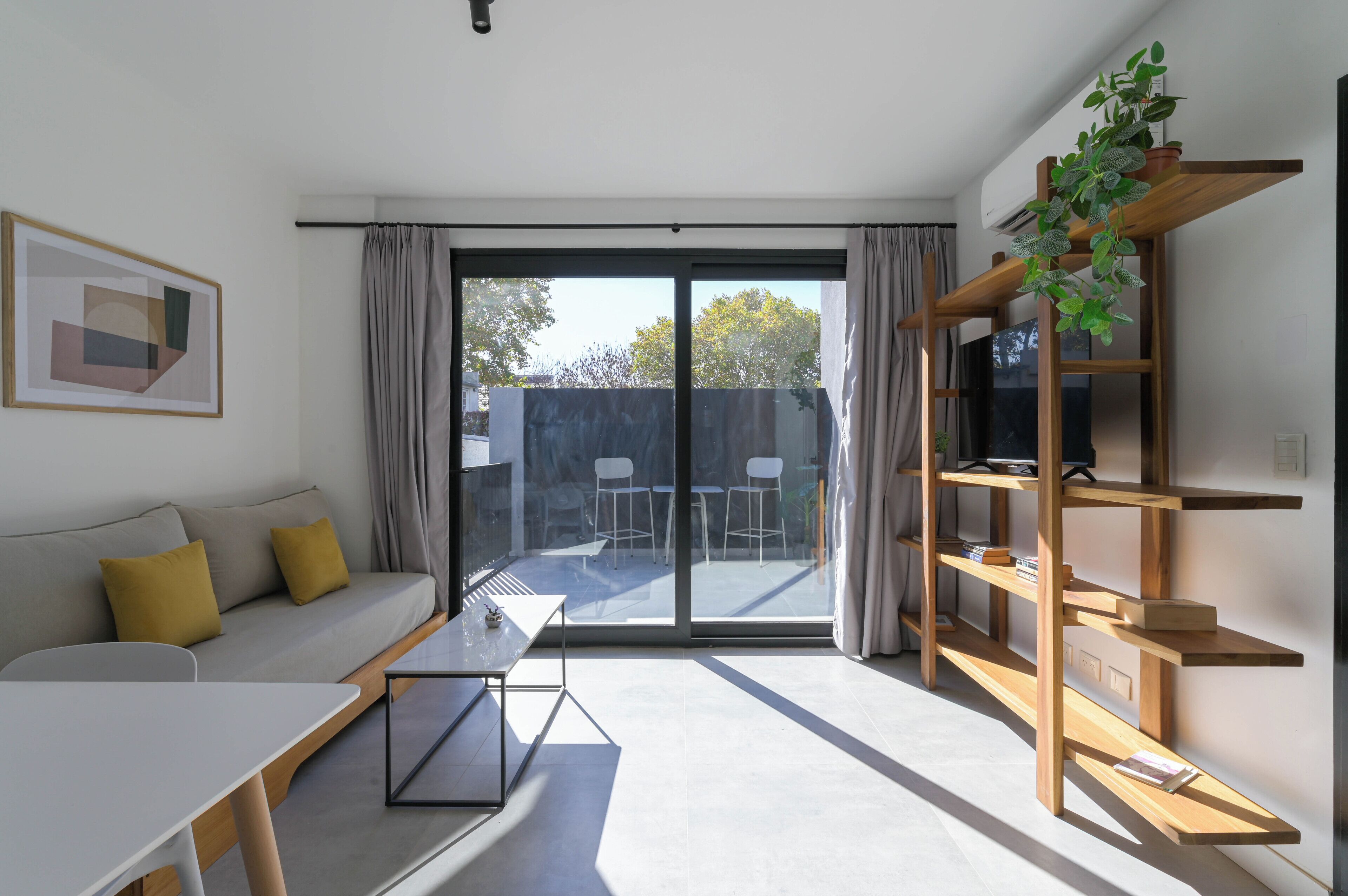 A modern living room with a sofa, a coffee table, a TV, and a sliding glass door leading to an outdoor area with chairs.