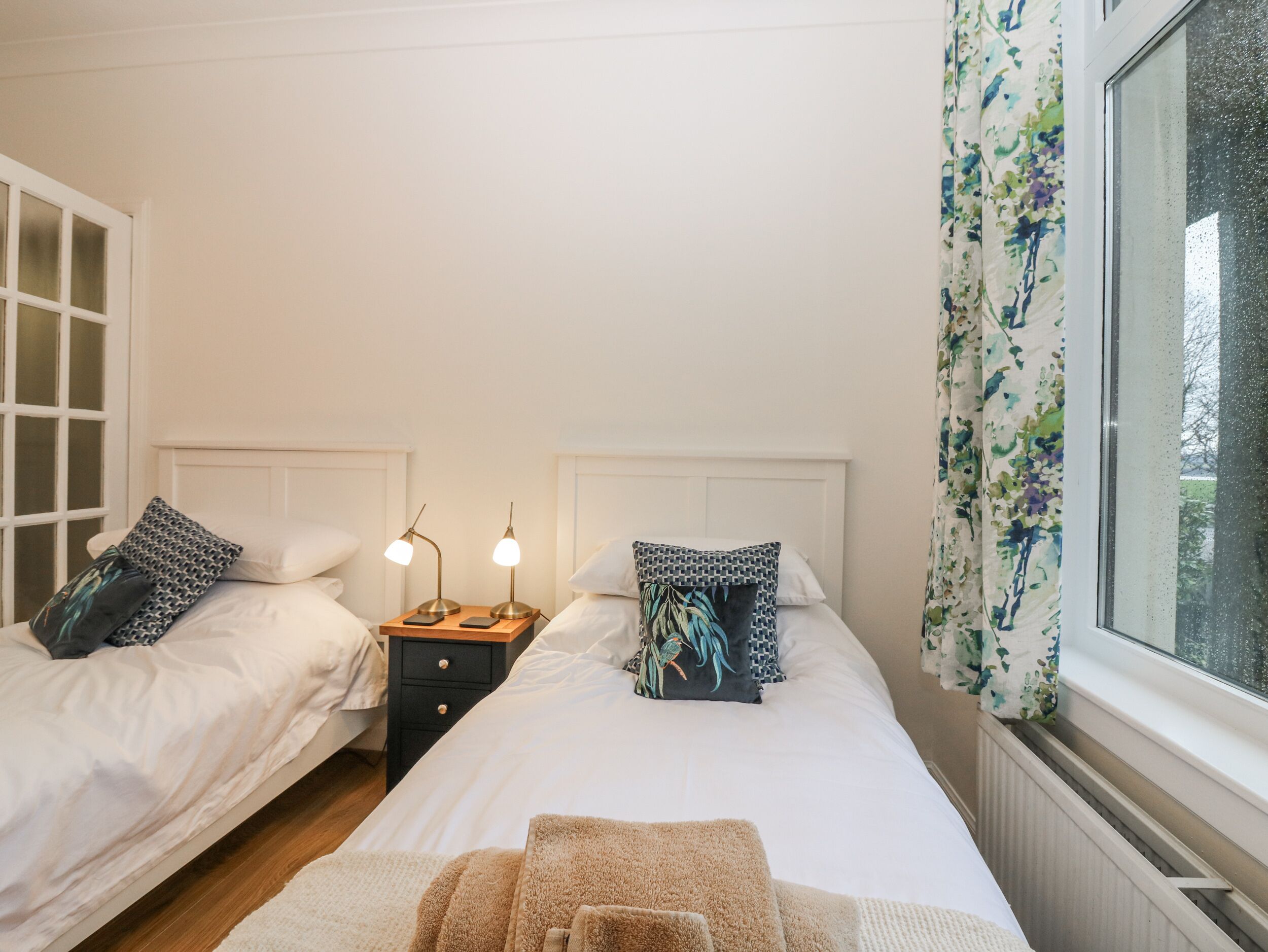 A bedroom with two beds, a nightstand with lamps, and a window with curtains.