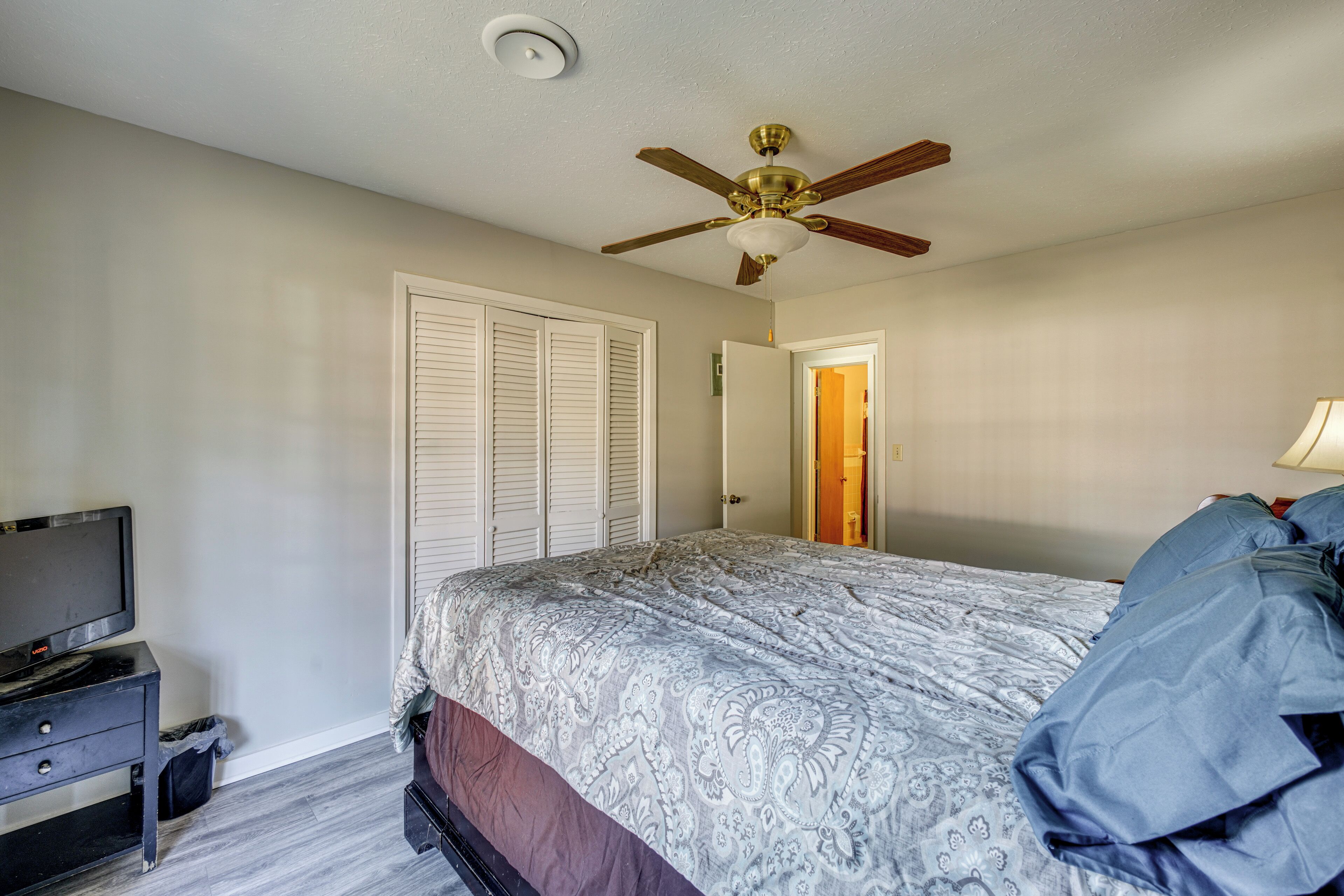 A bedroom with a bed, a ceiling fan, a television on a stand, and a door leading to another room.