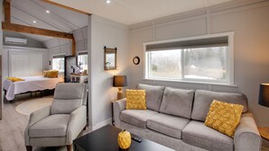 A modern living room with a grey sofa, armchairs, and a coffee table. There is a large window with a view of trees, a clock on the wall, and a lamp on a side table. - Bougie Barn 5 mi to Silverwood Theme Park! (Athol)