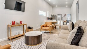 Ein modernes Wohnzimmer mit einem Flachbildfernseher, einem Holztische mit den Initialen 'M S', einem Ledersessel und einem gemusterten Teppich.