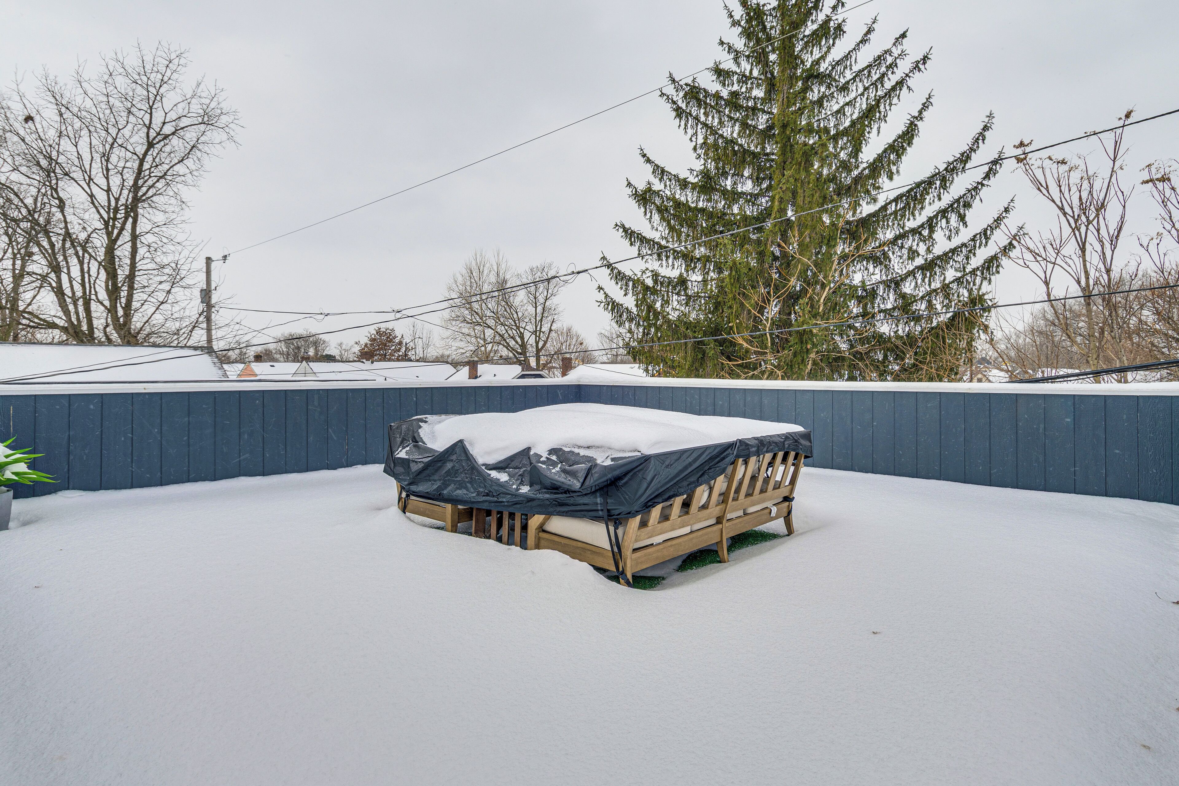 A covered outdoor seating area with a wooden bench and a table, surrounded by snow and trees.
