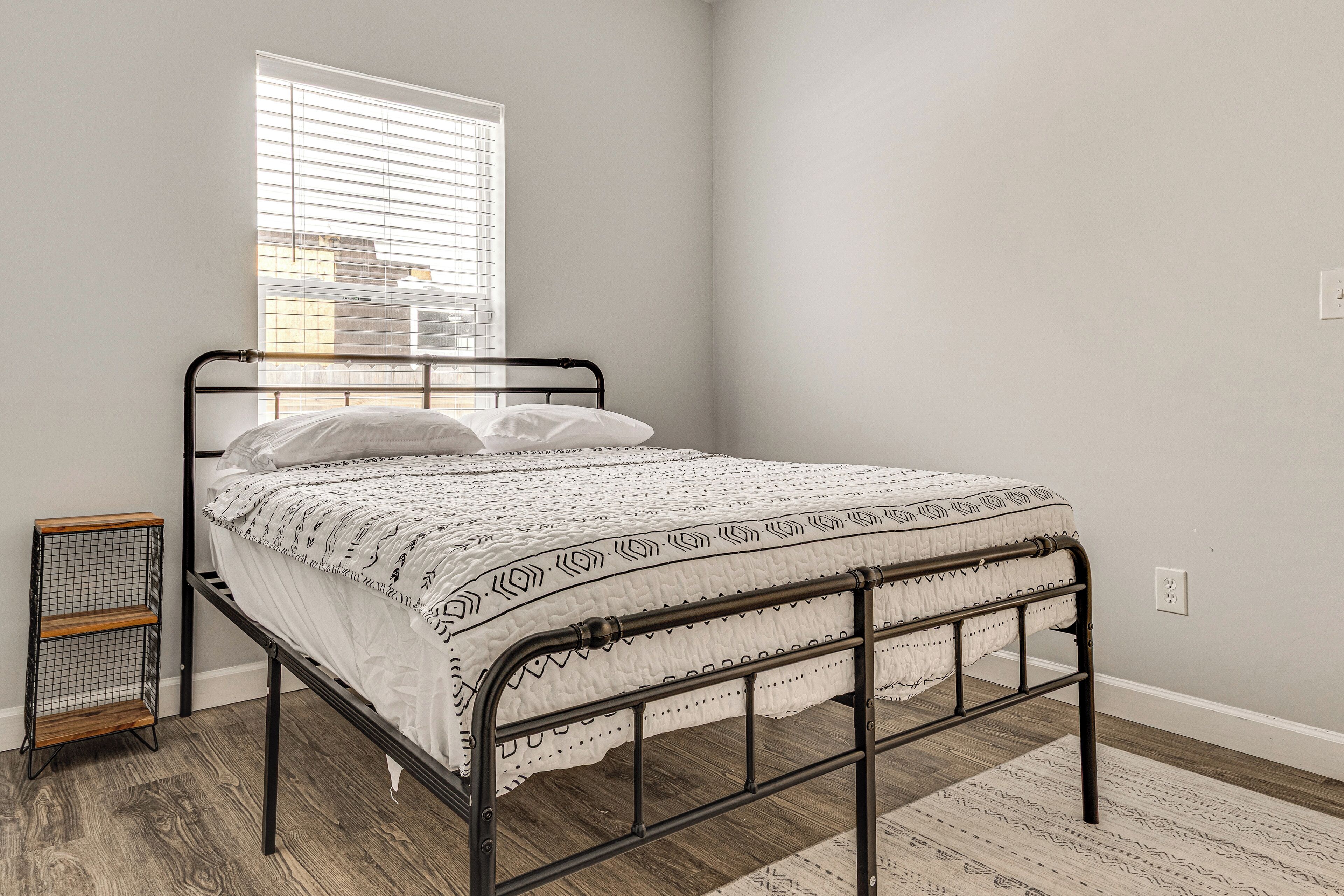 Una cama individual con estructura metálica, ropa blanca de cama y un edredón estampado. Un pequeño estante de madera está al lado de la cama, y se ve una ventana con persianas en el fondo.