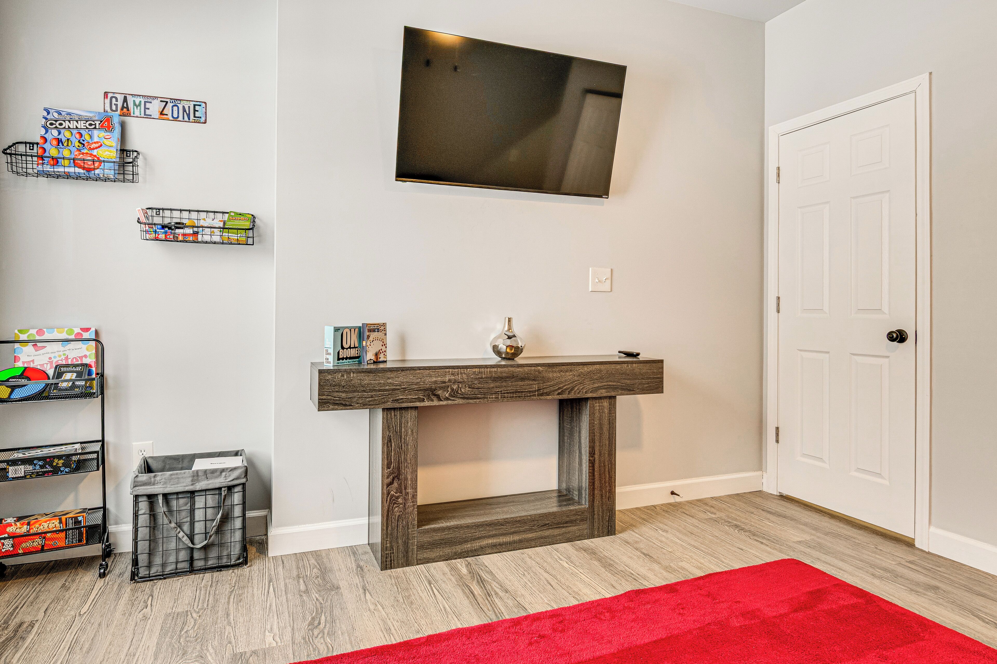 Een kamer met een houten console, een aan de muur bevestigde televisie, een witte deur en een rood vloerkleed.