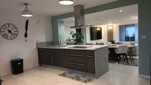 A modern kitchen with a central island, dark cabinetry, and pendant lighting. There is a dining area with a table and chairs, a large mirror, and a plant.