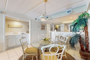 A dining area with a glass table, white wicker chairs, a central light fixture, and a kitchen in the background. - Shorefront Resort Retreat: Suntide III - Unit 1002 (South Padre Island)