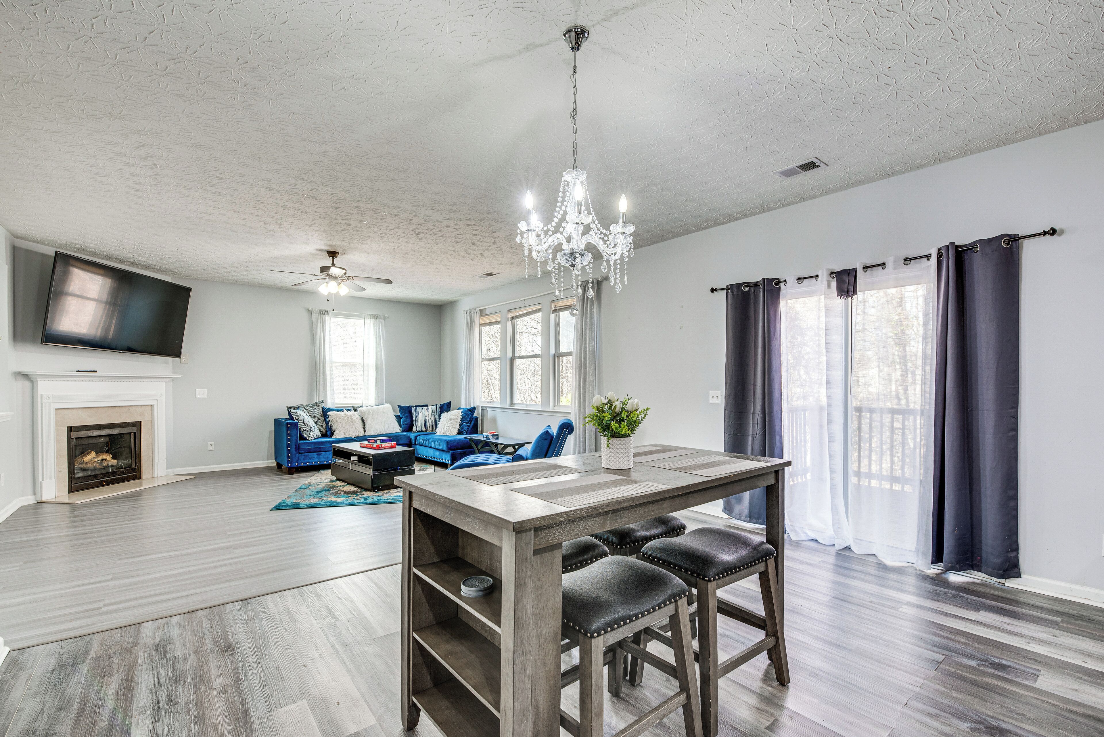 A spacious living room with a fireplace, a blue sofa set, a wooden dining table with bar stools, and a chandelier.