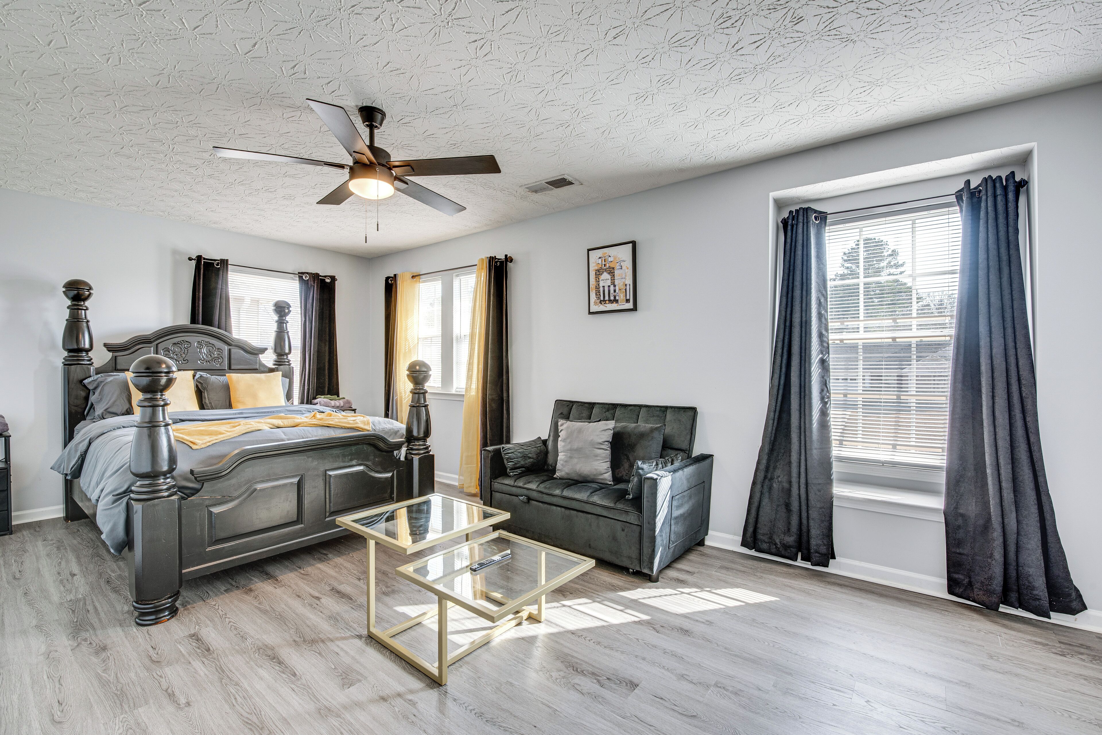 A bedroom with a bed, a sofa, a glass coffee table, a ceiling fan, and a window with curtains.