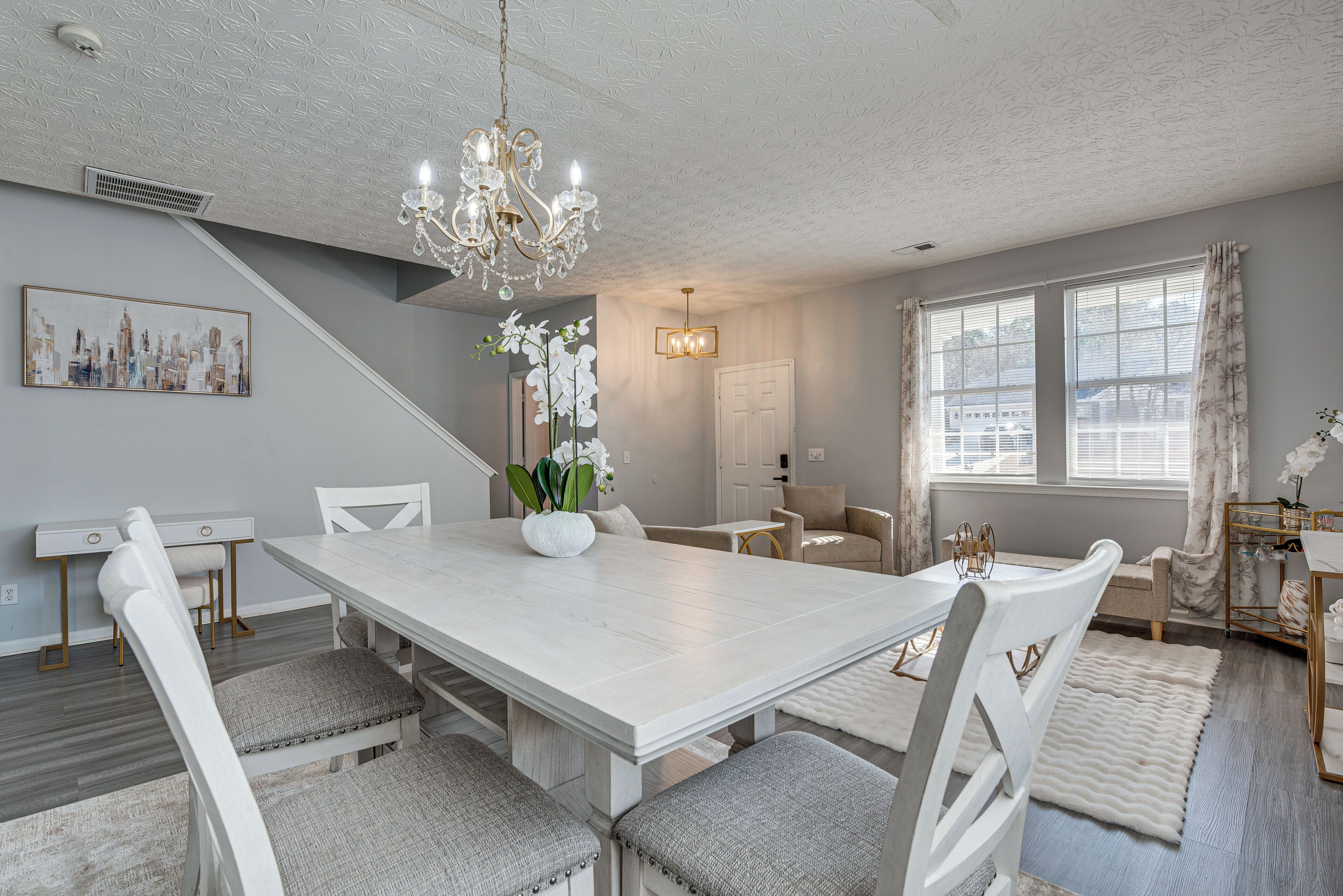 A dining area with a white table, grey upholstered chairs, a chandelier, and a cityscape painting on the wall.