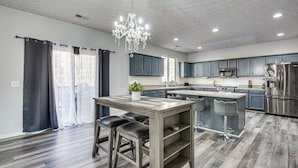 A modern kitchen with grey cabinets, a central island with bar stools, a chandelier, and a stainless steel refrigerator. - Great for Gatherings! Elegant Atlanta Retreat (Atlanta)