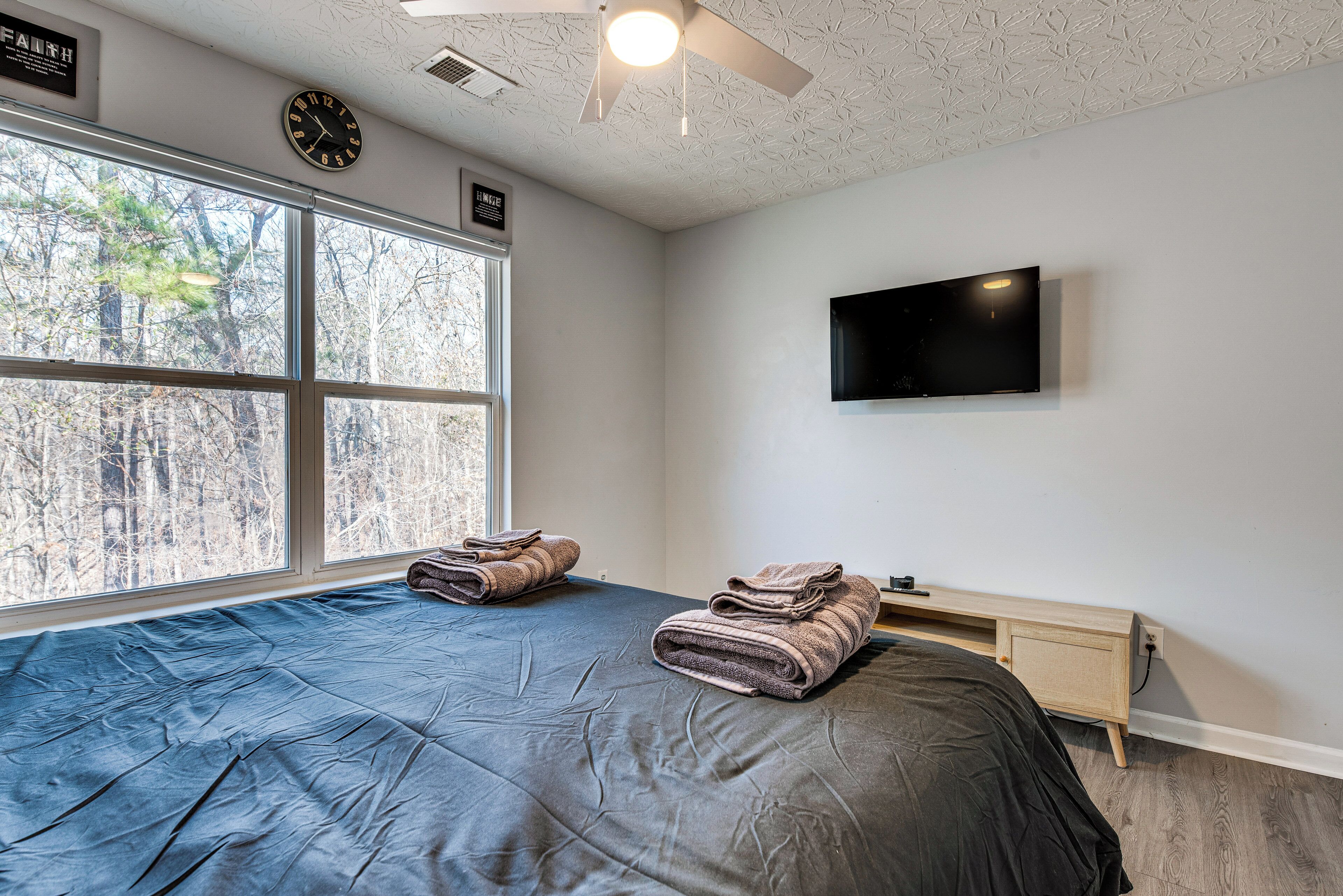 Une chambre avec une grande fenêtre, un lit recouvert d’une couverture foncée, un téléviseur à écran plat fixé au mur et un ventilateur de plafond.