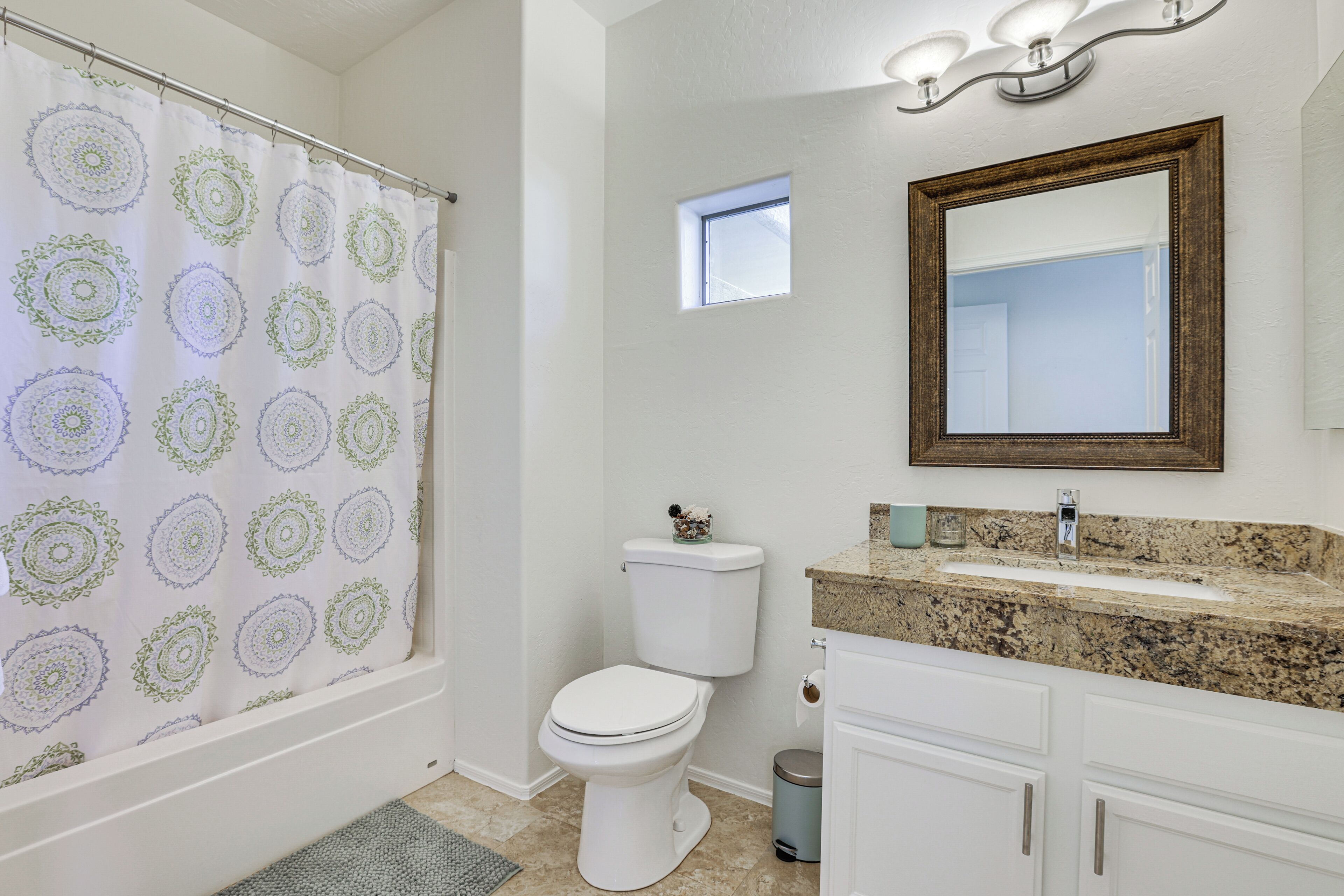 A bathroom with a shower curtain, toilet, sink, mirror, and a small window.