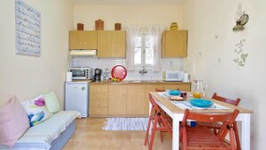 A compact kitchen with wooden cabinets, a dining table with chairs, a small refrigerator, and a window with curtains. - Naoussa Apricot Garden, 5' to Center, 5' to Beach (Naousa)