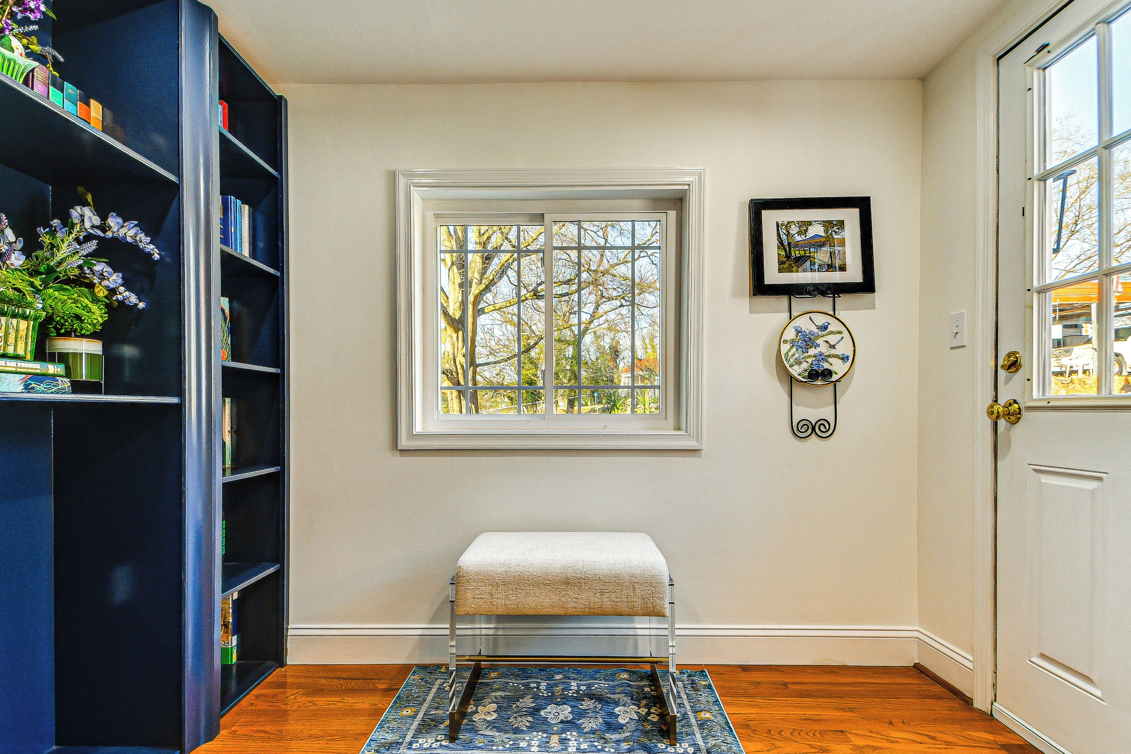 Una habitación con suelo de madera, un banco blanco, una estantería, una ventana con vistas y una puerta.