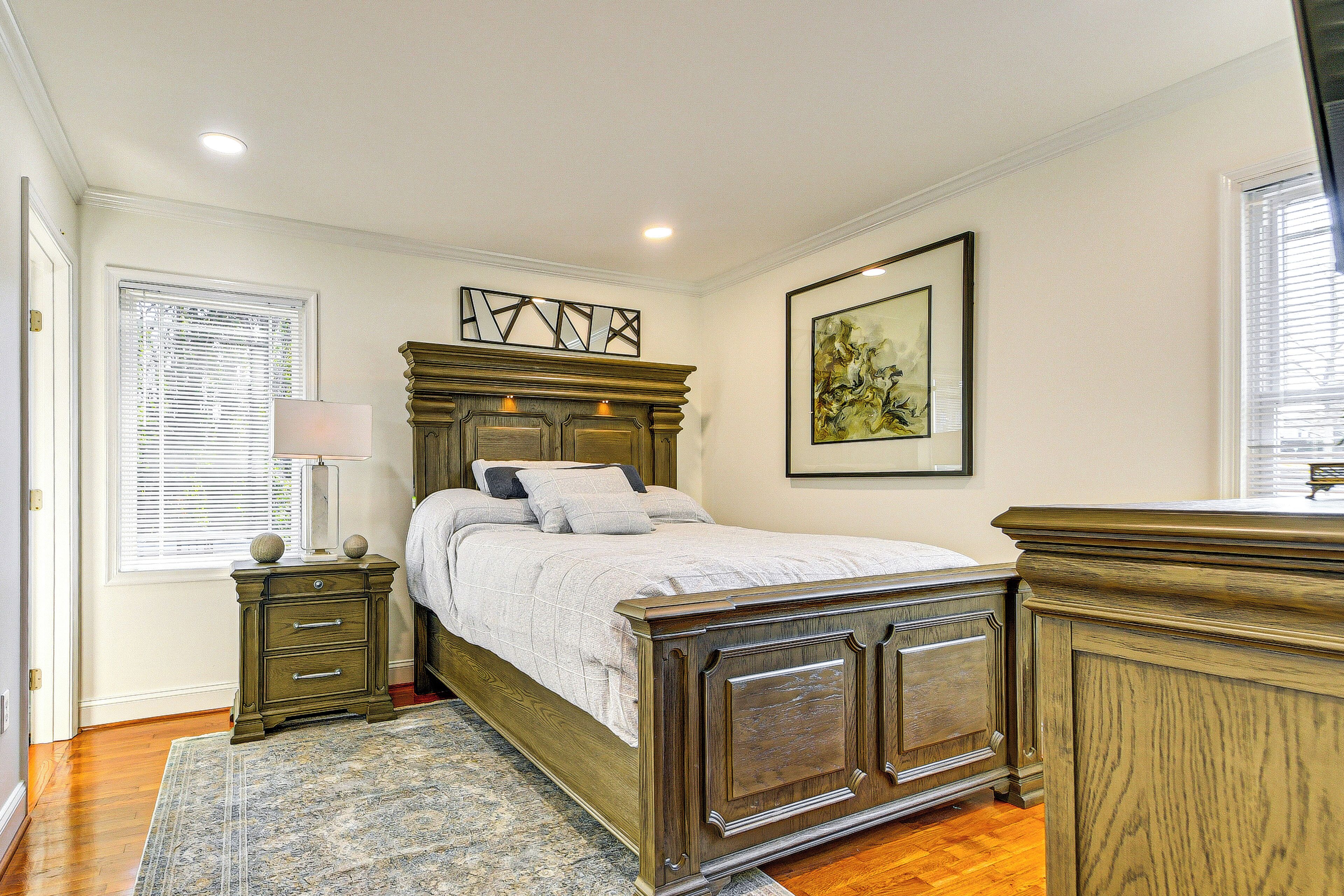 Un dormitorio con cama de madera, mesita de noche, cómoda, ventana con persianas e imagen enmarcada en la pared.