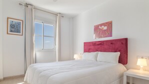 Une chambre à coucher avec un grand lit, deux tables de chevet avec des lampes, une fenêtre avec des rideaux et deux œuvres d’art murales.