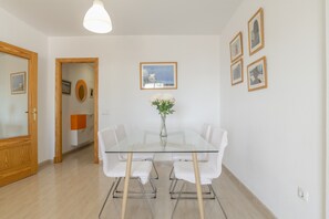 Un espace de salle à manger moderne avec une table en verre, des chaises blanches, un vase rempli de fleurs et des tableaux encadrés accrochés au mur.