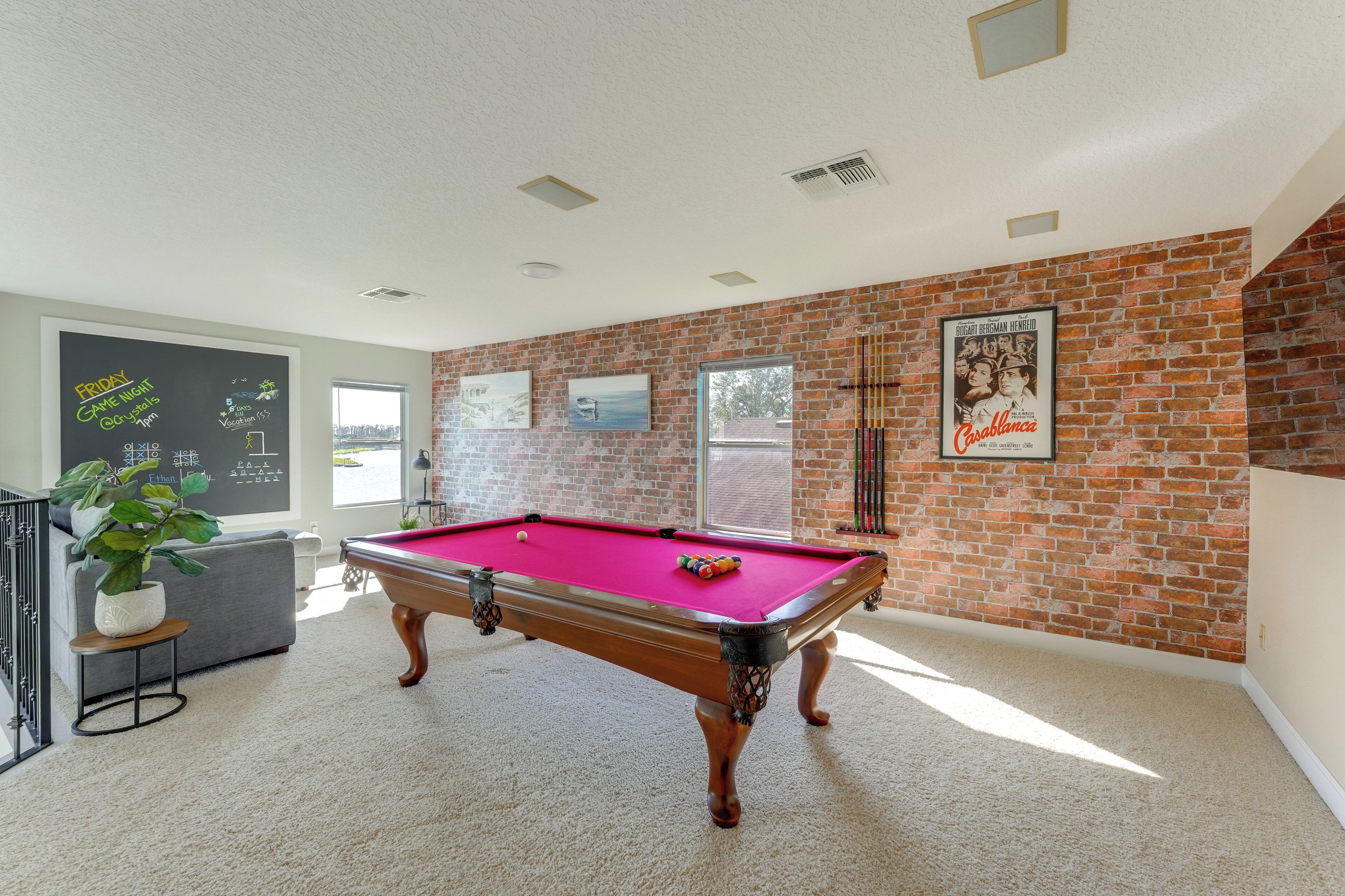 A pool table in a room with a brick wall, a chalkboard with writing, and a poster on the wall.