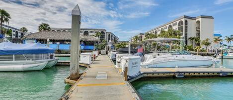 A marina with boats docked, a thatched-roof building, and a multi-story hotel in the background.