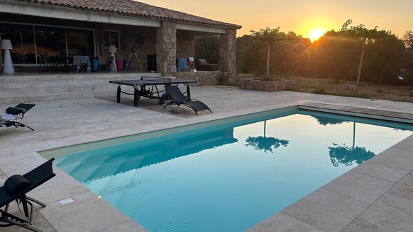 Ein Swimmingpool mit Liegestühlen und einem Tisch, umgeben von einem Steinhaus mit großen Fenstern.
