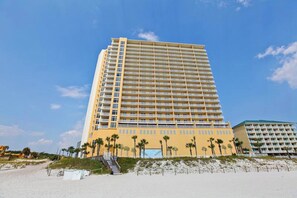 Un hotel alto de varios pisos, con techo plano y balcones, situado cerca de una playa con palmeras.