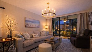 Ein modernes Wohnzimmer mit einer Couch, einem Hocker, einem Couchtisch und einer Deckenleuchte. An der Wand hängt ein Gemälde, ein Beistelltisch mit einer Lampe und ein Blick ins Freie durch große Fenster.