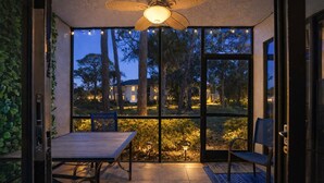 Eine Terrasse mit Tisch und Stühlen, einem Deckenventilator und Blick auf Bäume und Gebäude im Dämmerlicht.