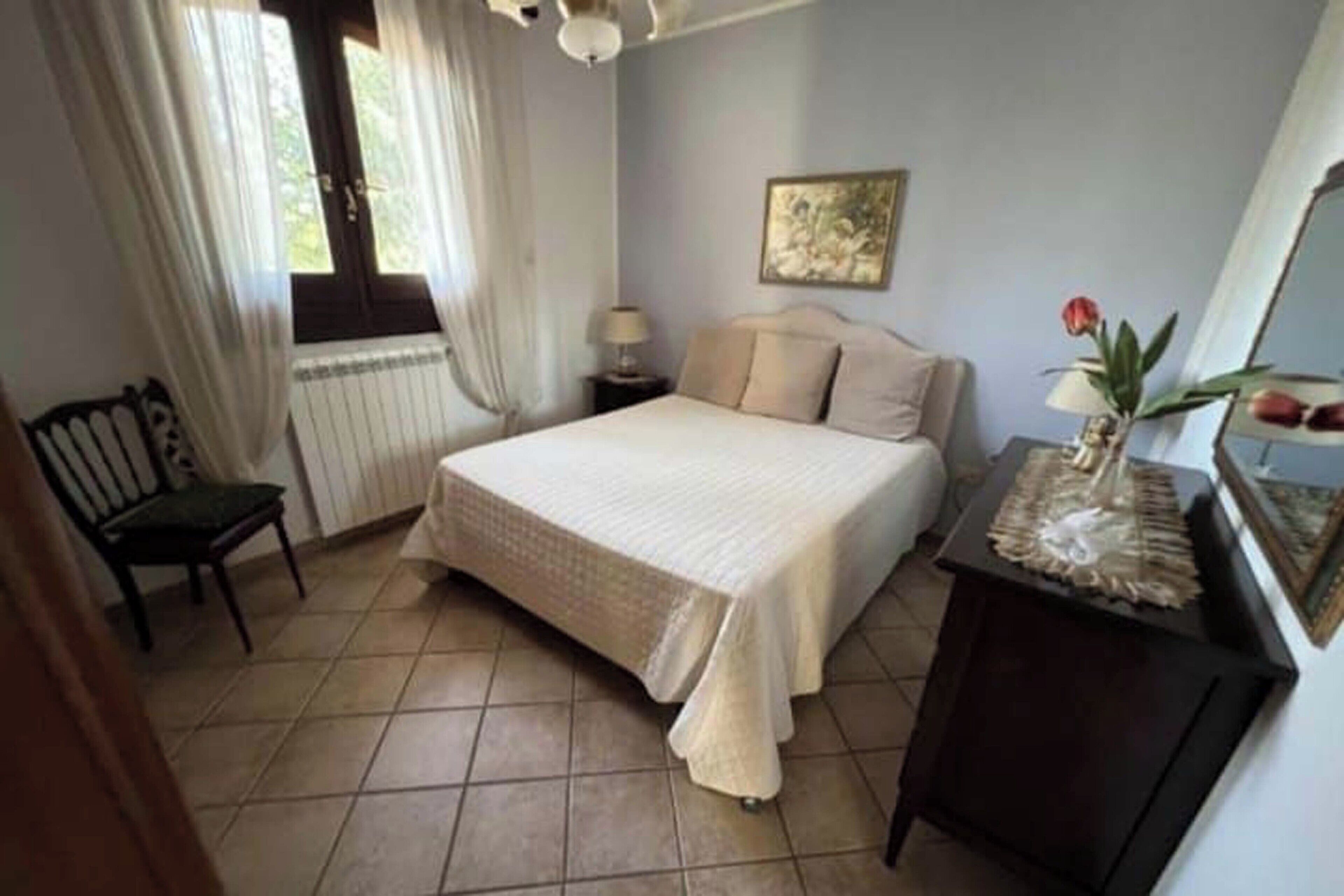 Een slaapkamer met een bed, een stoel, een nachtkastje, een lamp, een spiegel en een raam met gordijnen.